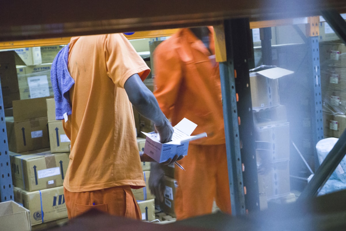 Two warehouse workers in orange uniforms inspecting a box against paperwork beside stocked shelving