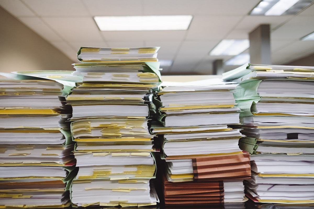 Tall stacks of paper documents and folders with yellow tabs piled on an office desk