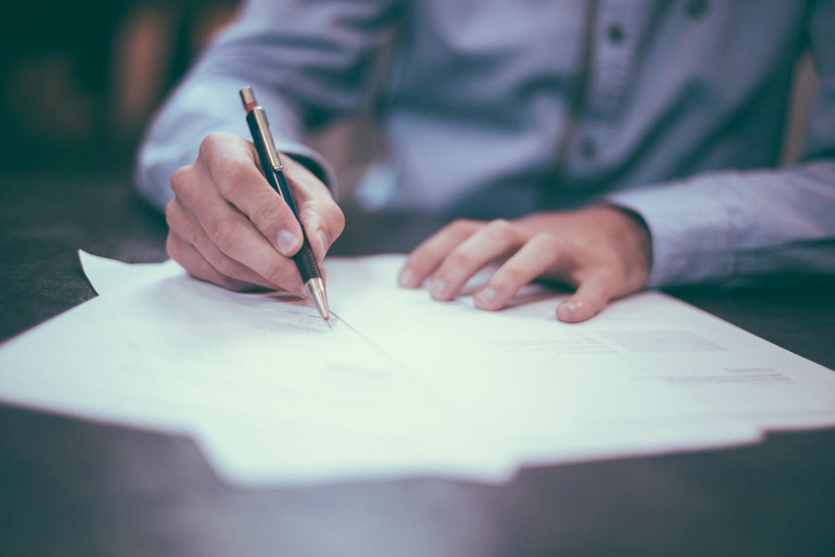 Close-up of a person in a light blue shirt signing a paper document with a pen at a desk