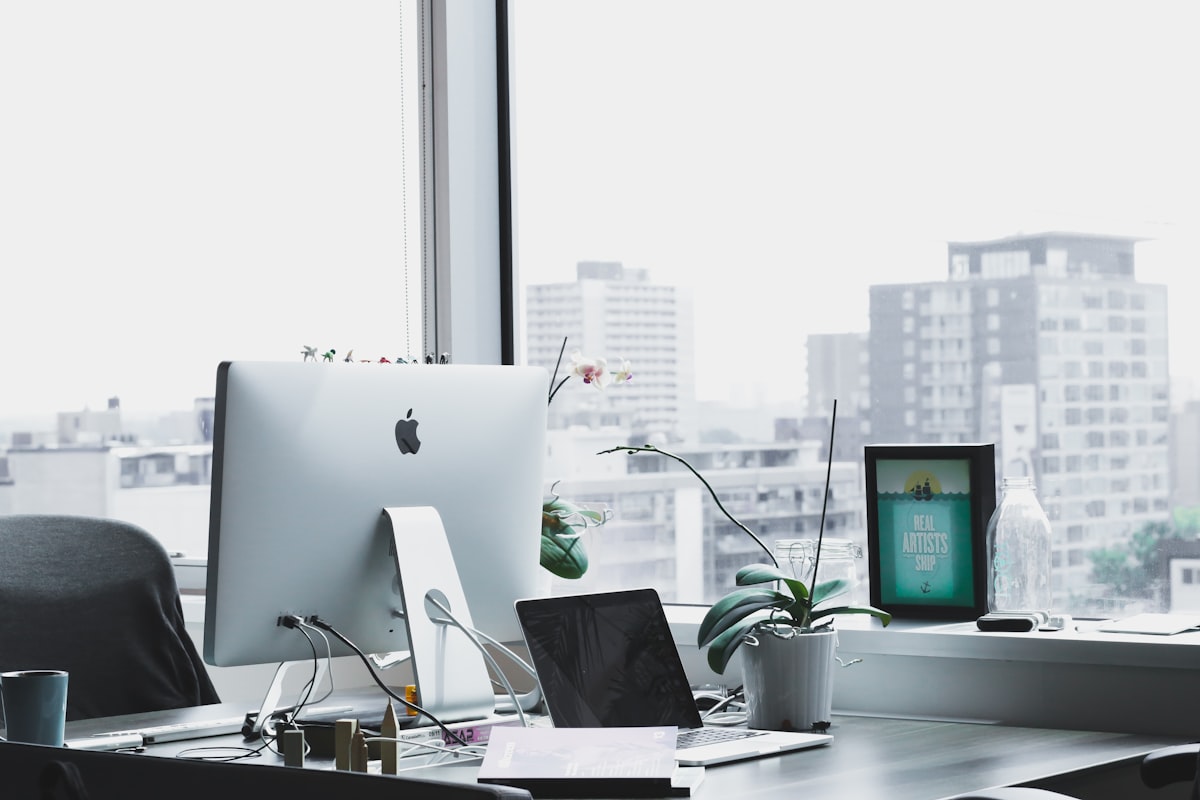 A desktop computer and laptop with accessories on a bright office desk beside a window