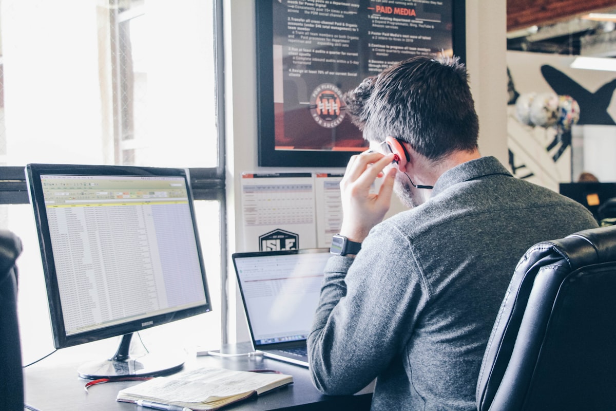 A man at a desk working on a desktop monitor showing a spreadsheet while holding a phone